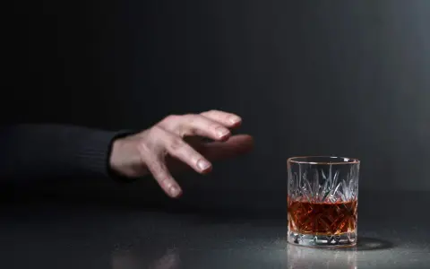 Mano tesa verso un bicchiere di whisky, un'immagine che evoca la domanda: quanto beve un alcolista al giorno?