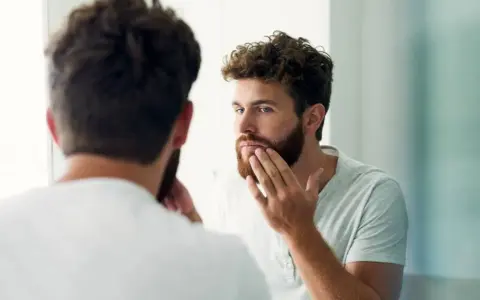 Hombre con barba regia se mira al espejo, acariciando su rostro.