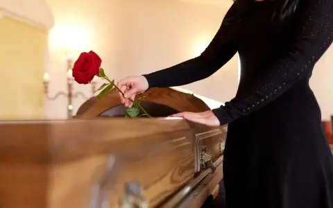Kobieta w czarnej sukni kładzie czerwoną różę na trumnie. W tle widać świeczniki, przypominające o przesądach pogrzebowych i wieńcach.