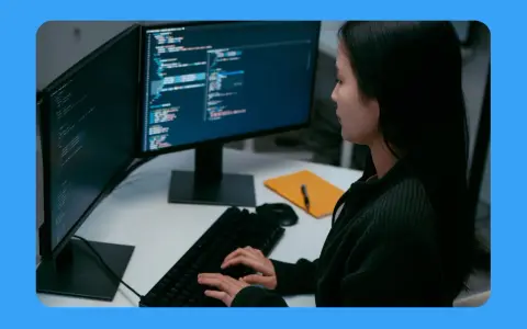 Jeune femme travaillant sur un ordinateur, avec deux écrans affichant du code. Sa position devant l'écran du bureau est concentrée.