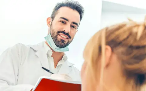 Uśmiechnięty dentysta w białym fartuchu i masce na szyi rozmawia z pacjentką, trzymając notatnik.