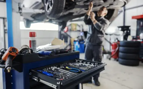 Warsztat samochodowy: mechanik pracuje pod podniesionym autem, wózek z narzędziami na pierwszym planie.