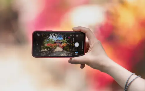Une main tient un smartphone pour capturer un escalier fleuri menant à un pavillon. L'écran montre les bases de la photographie, avec des options comme "PHOTO" et "PORTRAIT".