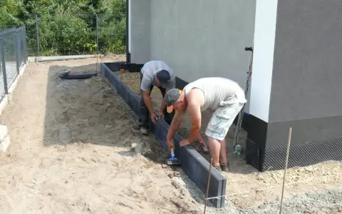 Dwóch mężczyzn pracuje nad przygotowaniem podłoża pod kostkę, układając obrzeża.