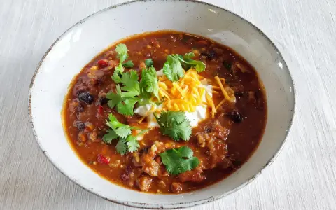 Gęste, aromatyczne chili con carne z fasolą, mięsem i przyprawami, udekorowane śmietaną, startym serem i świeżą kolendrą.