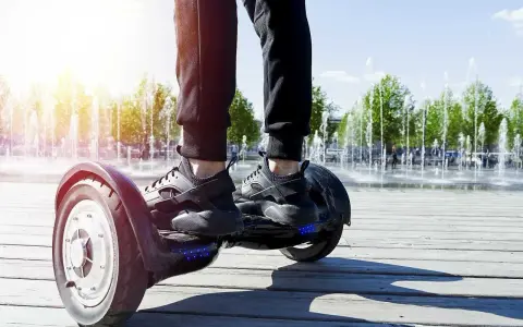 Osoba na deskorolce elektrycznej, czyli urządzeniu transportu osobistego, porusza się po drewnianej promenadzie. W tle widać fontanny i drzewa.