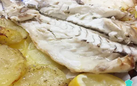 Lubina al horno jugosa y tierna, acompañada de patatas asadas y rodajas de limón fresco.