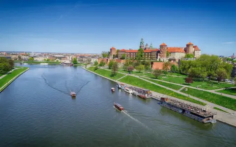 Widok na Wawel i Wisłę. Kilka łodzi pływa po rzece, a na brzegu zielony park i zabudowania.