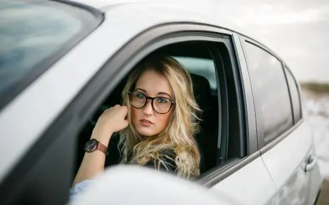 Blondynka w okularach siedzi za kierownicą białego samochodu. Pokazuje, jak nosić okulary, by wyglądać stylowo i pewnie.