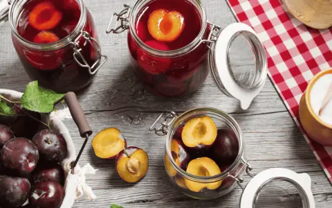 Frische Pflaumen werden für das Einkochen vorbereitet. Ganze und halbierte Früchte liegen neben Gläsern mit eingelegten Pflaumen.
