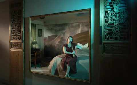 Elisa Miller sits on a bed, her reflection captured in a large mirror. The room features ornate wooden panels and a dramatic mountain mural.