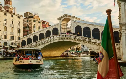 Un vaporetto naviga sul Canal Grande, passando sotto il Ponte di Rialto. L'Italia è rappresentata da una bandiera sventolante.