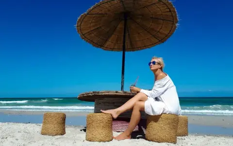 Kobieta w białej sukience relaksuje się pod parasolem na plaży, popijając napój. Idealne tunezja wakacje.