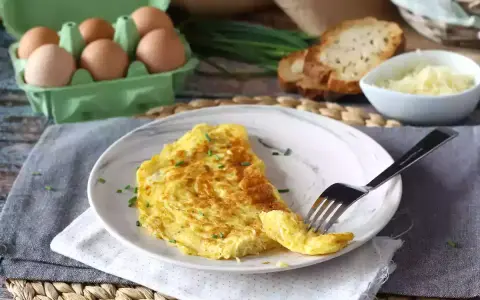 En gyllene omelett med ost, toppad med gräslök. Bredvid syns ägg, rostat bröd och mer ost. En perfekt start på dagen!