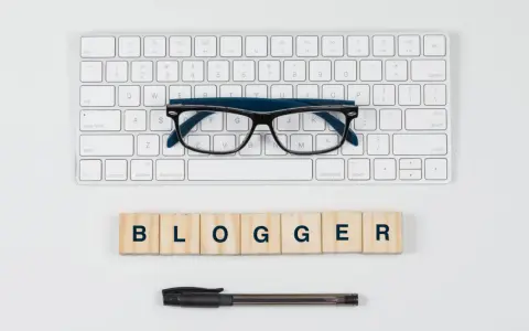 Okulary, klawiatura i drewniane klocki z napisem "BLOGGER" - idealne narzędzia do pisania bloga po angielsku.