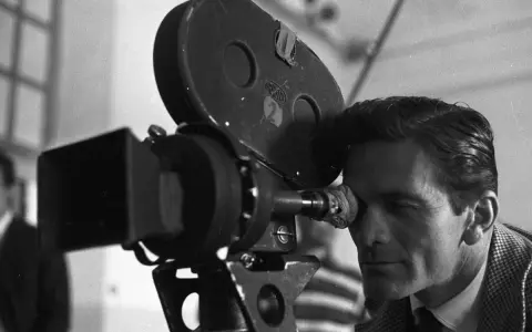 Pier Paolo Pasolini, con lo sguardo concentrato, osserva attraverso l'obiettivo di una cinepresa Arriflex, pronto a catturare la realtà.