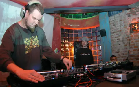 DJ Maceo Wyro w akcji, miksuje muzykę w klubie. W tle widać innych DJ-ów i kolorowe oświetlenie.