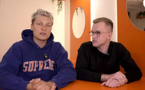 Dwóch mężczyzn siedzi przy pomarańczowym stole. Jeden w niebieskiej bluzie z napisem "SUPREME", drugi w czarnej koszulce. Rozmawiają o kebabie friza.
