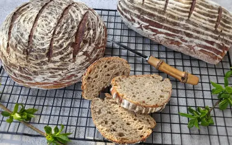 Dwa bochenki chleba żytniego, jeden okrągły, drugi podłużny, leżą na kratce. Obok pokrojone kromki, nóż z drewnianą rączką i zielone gałązki.