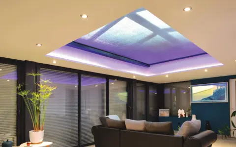 Salon moderne avec extension de maison et son puits de lumière illuminé de violet. Canapé confortable, plantes vertes et grandes baies vitrées.