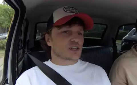 Mężczyzna w czapce z daszkiem i białej koszulce, siedzący w samochodzie. W tle widać drzewa i inne pojazdy. To może być początek jego zaorski wiek.