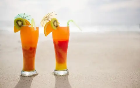 Dwa drinki Malibu z sokiem pomarańczowym, ozdobione plasterkami kiwi i pomarańczy, stoją na piaszczystej plaży.