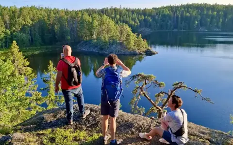 Vandring i Tyresta nationalpark. Tre personer njuter av utsikten över en stilla sjö och en tät skog. En man tar foto, en annan spanar ut, och en tredje vilar på en klippa.