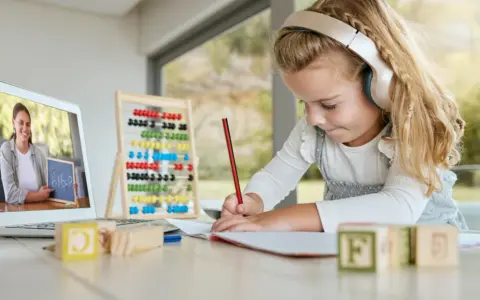 Dziewczynka w słuchawkach uczy się matematyki online, pisząc w zeszycie. Na ekranie laptopa nauczycielka pokazuje zadanie.