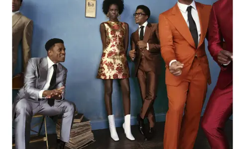 A group of stylish individuals in smart cocktail attire, featuring bold suits and a patterned dress, pose against a blue wall.