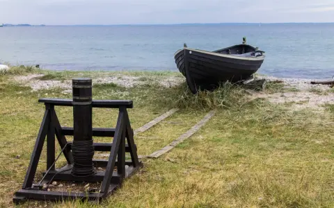 A dark wooden boat rests on a grassy shore next to a winch and wooden tracks leading to the sea.