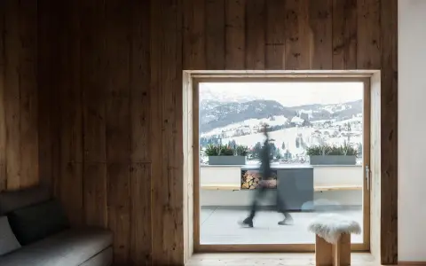 Interni di case di montagna con pareti in legno scuro, travi a vista e un divano grigio. Fuori, un paesaggio innevato.