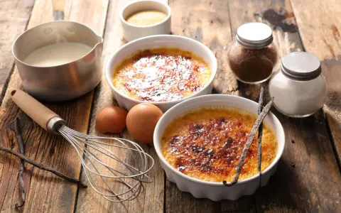 Dwa kremowe desery creme brulee z karmelizowanym cukrem, obok składniki: śmietanka, jajka, laski wanilii, cukier.