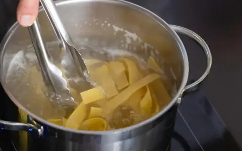 Szczypce do makaronu wkładają szerokie wstążki makaronu pełnoziarnistego do gotującej się wody w garnku.