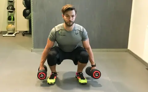 Mann macht Kniebeugen mit Kurzhanteln. Er hält zwei Hanteln mit je 14 kg Gewicht.