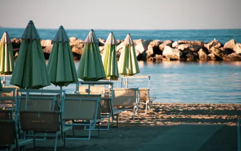 Ombrelloni verdi e lettini vuoti sulla spiaggia di una città riviera romagnola, con il mare calmo e una diga di sassi sullo sfondo.