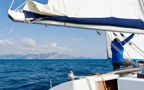 Mann im blauen Anzug stellt das **Rigg segeln** ein, während das Boot über das blaue Meer gleitet. Berge im Hintergrund.