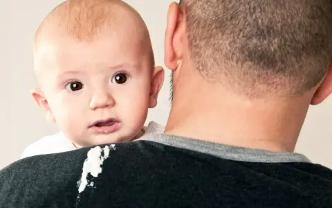 En söt bebis med stora ögon tittar fram över pappans axel. En liten vit fläck syns på pappans tröja, kanske från en rapning.