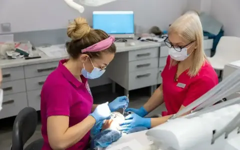 Dwie dentystki w maseczkach i rękawiczkach pracują przy pacjencie. Jedna z nich używa narzędzi, druga asystuje. Zabieg bez bólu dzięki lidokainie.