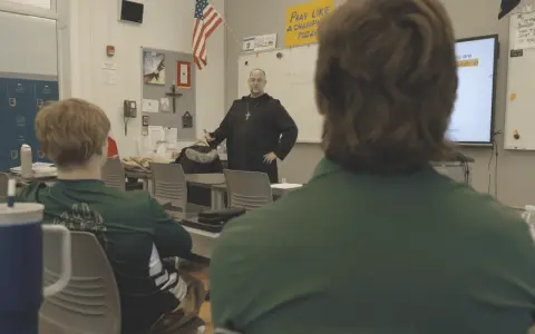 A monastery leader in black robes addresses students in a classroom.