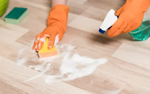 Hands in orange gloves spray cleaner and scrub a vinyl plank floor with a sponge, showing how to clean vinyl plank flooring.