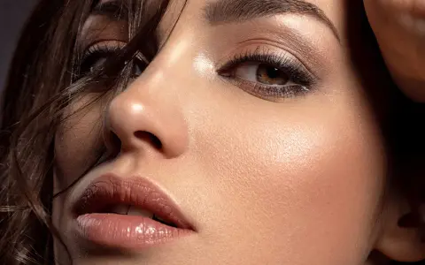 Primo piano di un volto femminile con un sofisticato trucco occhi marroni, labbra luminose e pelle radiosa.