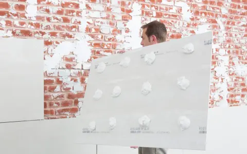 Mann hält Gipskartonplatten mit Klebepunkten, um sie auf unebener Wand zu befestigen.