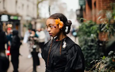 Ragazza con trecce davanti e capelli sciolti, ornata da un fiore arancione e makeup audace. Indossa un abito nero con dettagli voluminosi.