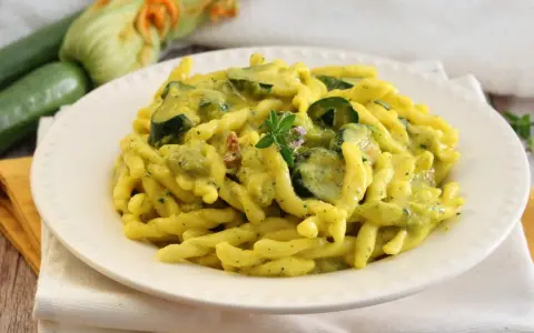 Piatto di pasta con zucchine e zafferano, guarnito con un rametto di erbe aromatiche. Sullo sfondo zucchine fresche e fiori di zucca.
