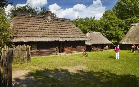 Skansen Chorzów bilety: drewniane chaty kryte strzechą, dzieci bawią się na trawie, zwiedzający podziwiają architekturę.