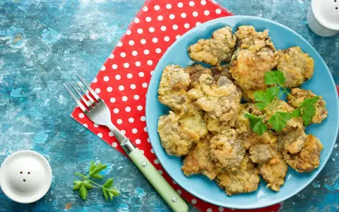 Piatto di fegatini di pollo impanati e fritti, guarniti con prezzemolo. Accanto, una forchetta e un sale e pepe.