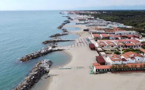 Vacker vy över pisa strand med sina stenpirar som bryter vågorna. Längre bort syns hus och en grönskande skog.