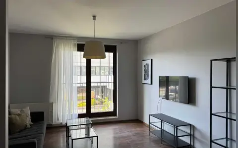 Nowoczesny salon z szarą kanapą, szklanymi stolikami i telewizorem. Widok z okna na zielony dziedziniec, idealne miejsce na relaks w śródmieście Wrocław.