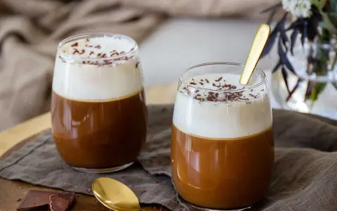 Två glas med krämig kaffe Karlsson, toppad med vispgrädde och chokladströssel. En gyllene sked vilar i det ena glaset.