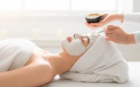 A spa technician applies a white mask to a woman's face. She's wrapped in a towel, enjoying a relaxing facial or massage first.
